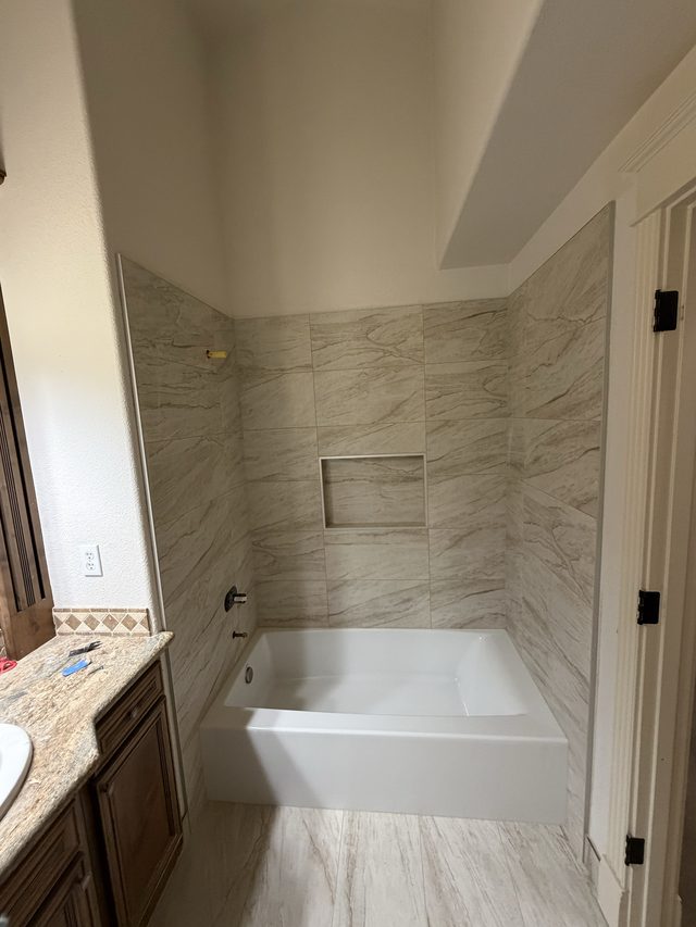 alcove bathtub with beige stone-look tile surround, niche, vanity at left